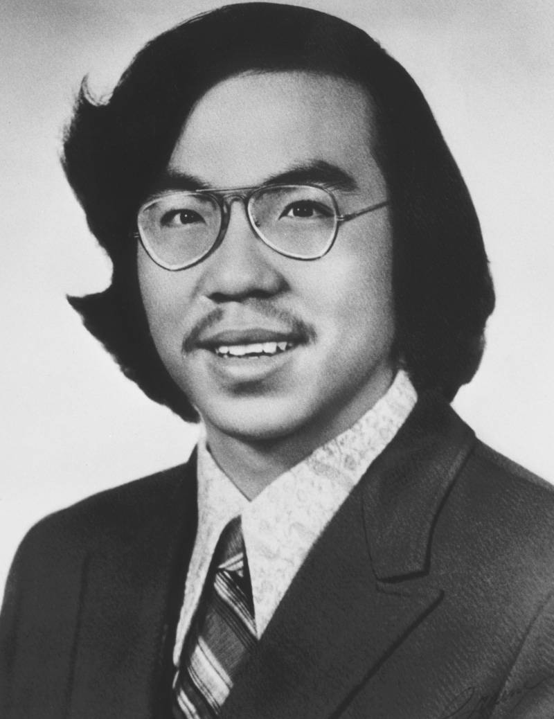 A portrait of a man with long hair, wearing a tie. The photo is in black and white. He is smiling.