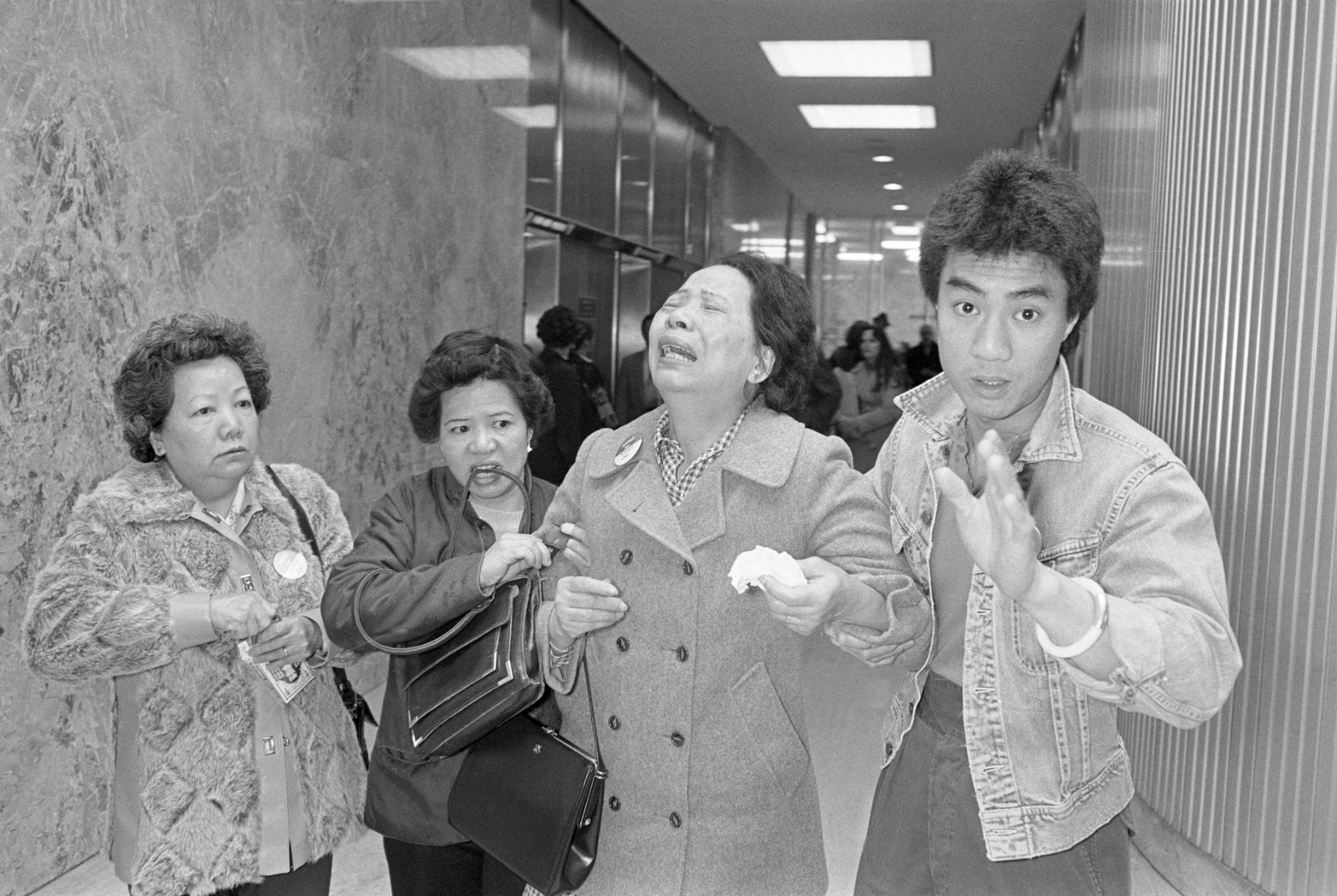 Three women, left, and one man, right, walk down a hallway in a black and white photo. A woman third form left is crying in anguish, as the woman amd man to her immediate sides carry her by the arms.
