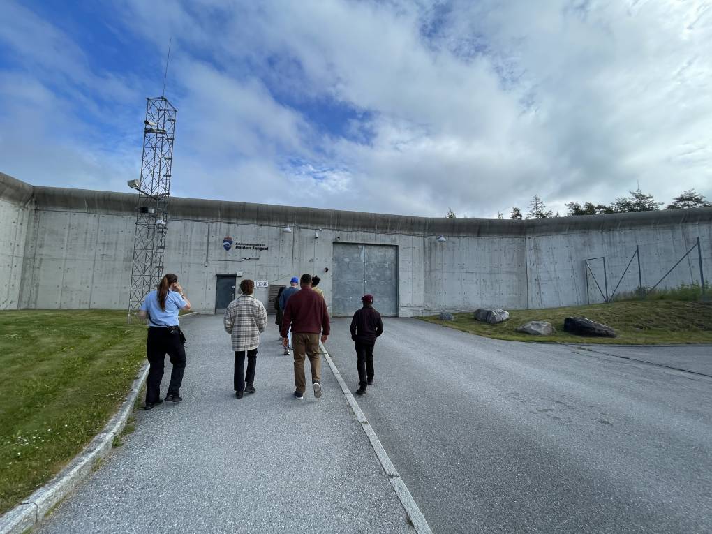 A small group of people stand outside a prison facility in Norway. They're facing a large gray wall which looks like concrete.