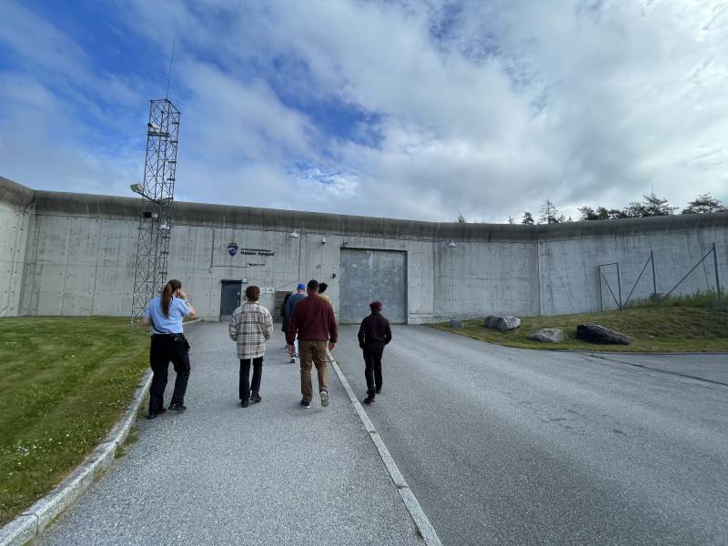 A small group of people stand outside a prison facility in Norway. They're facing a large gray wall which looks like concrete.