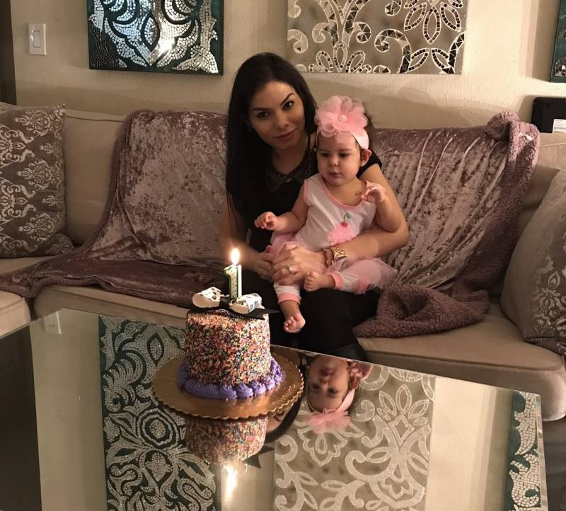 A woman holds a baby while sitting on a couch in front of a cake with a lit candle on top.