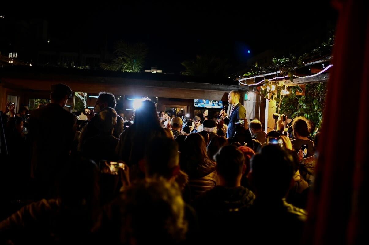 A shadowy photo showing a crowd of supporters surrounding Chesa Boudin, who is standing on a beer keg to address the crowd outside at a restaurant.