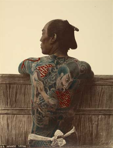 A Japanese man stands shirtless with his back to the camera, leaning on a fence.  His entire back is tattooed in an intricate and colorful pattern.