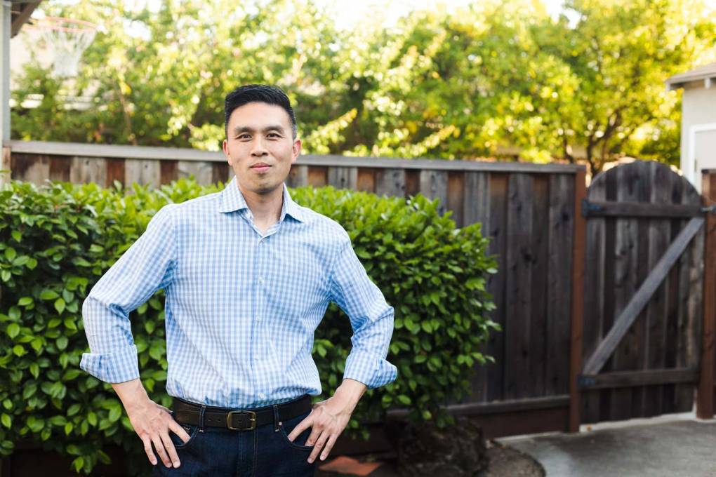 A light-skinned, trim Asian man in his late 30s or early 40s stands with his hands on his hips in front of a green hedge lining a wooden fence and a wooden gate, as if in a backyard, beyond which sunlight on green tree leaves is visible. He wears a blue-and-white checked collared shirt with the cuffs unbuttoned and neck undone, a white undershirt peeking out at the neck, dark jeans, and a belt. He is smiling confidently with his mouth closed and looking at the camera.