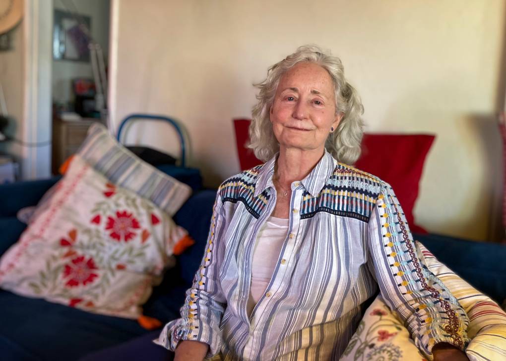 An older woman with peach skin and white hair sits on a blue couch in her living room, with red and blue pillows. She has blue eyes and a slight smile. She's wearing a blue and white striped shirt with geometric embroidery in yellow, blue, and rust along the shoulders and arms.