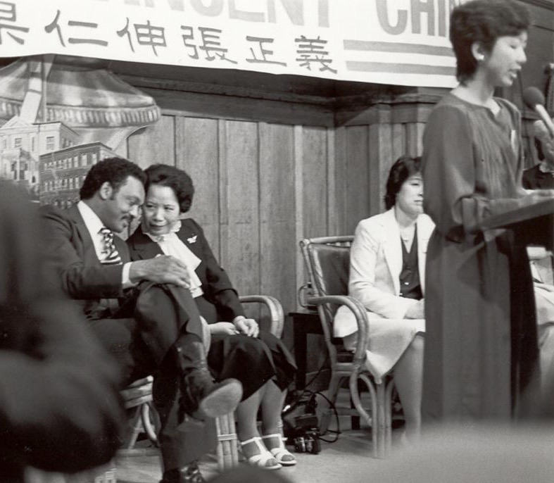 A black and white photo of people seated on a stage with a protest banner hanging behind them. 