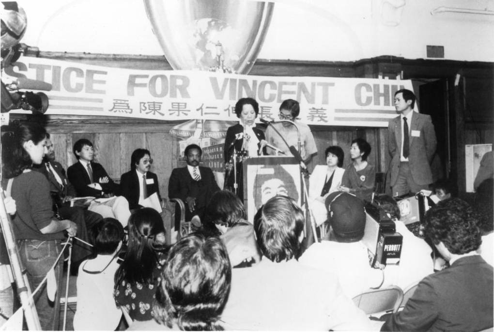 People are seated at a small stage in a black and white photo with "Justice for Vincent Chin" on a sign in English and Chinese in the background, a woman and man stand at a podium.