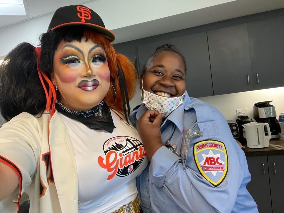 Panda Dulce in elaborate drag makeup, an SF Giants black and orange cap, and a white tshirt, posing for a selfie with a security guard in uniform. 