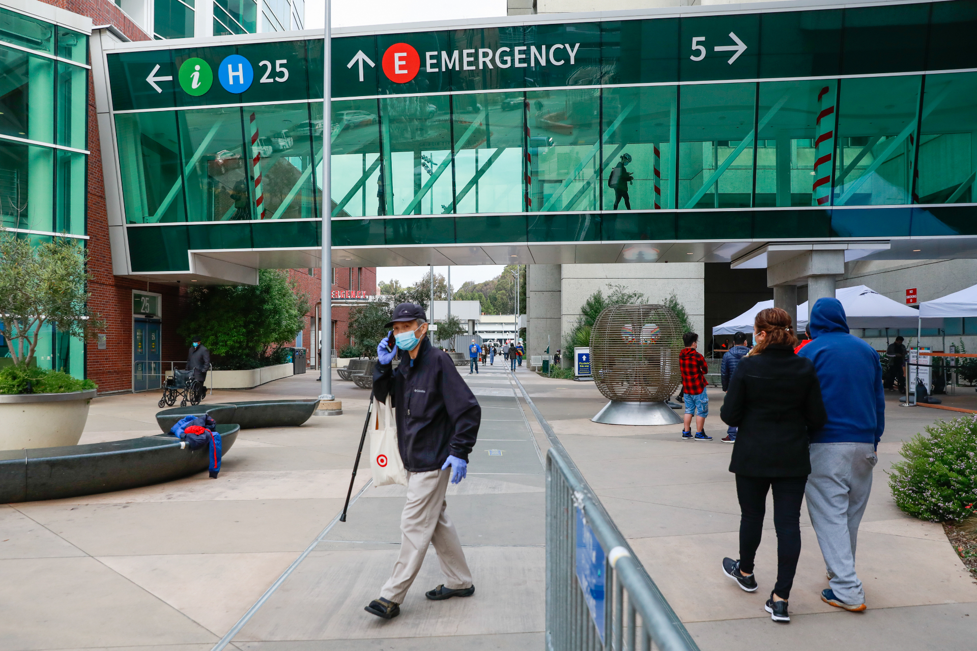 A hospital walkway is seen at the top of the frame as people walk through the area in front of San Francisco General Hospital in the foreground.