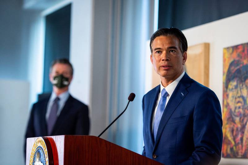 A fit, middle-aged Filipino man, with black hair slicked away from his head, stands at a wooden lectern inside a room, speaking toward, but beyond, the camera at his left. We see a sliver of the Seal of California at the top of the front of the lectern, and a skinny microphone neck extending from the lectern toward him. He wears a dark blue suit jacket, a white dress shirt, and a glossy powder blue tie. On a wall behind him are two paintings; the one visible behind him seems to be an oil or acrylic portrait in blues, pinks, and yellows or a man wearing a baseball cap and jacket, with a surprised or distraught look on his face. A tall man also dressed in a suit and wearing a black face mask stands at a distance in a doorway to Bonta's right; it appears to be Gov. Gavin Newsom.