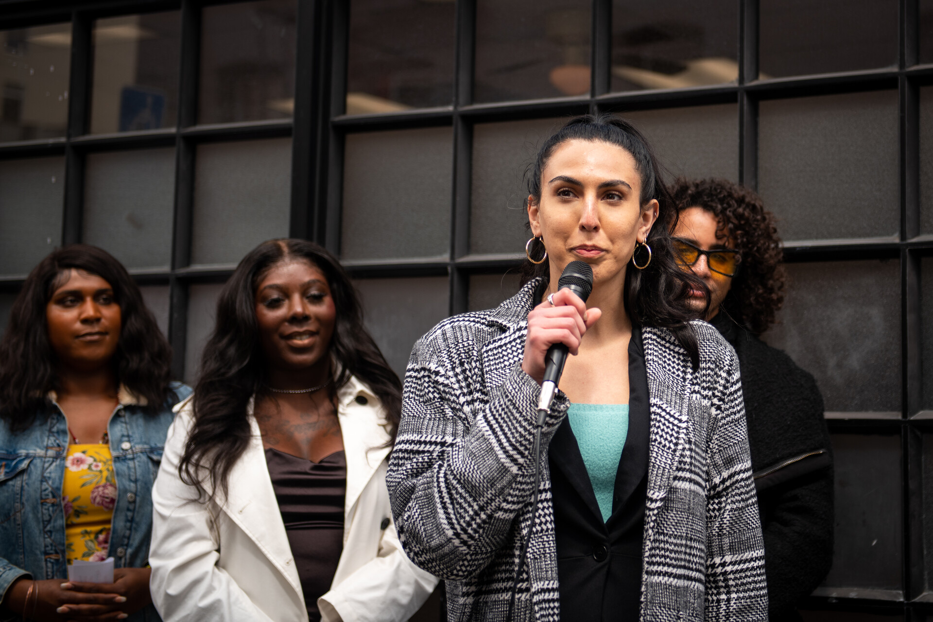 woman speaks into microphone she's holding on sidewalk outside building as three Black and brown people listen in the background
