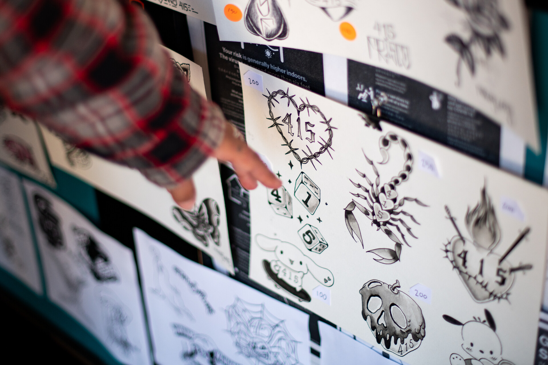 A hand points to sheets of tattoo designs hanging on the wall.  The designs feature 415 in honor of 415 Day.