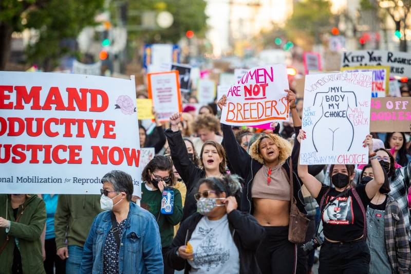 A large crowd moves through a San Francisco street. Many carry signs that read "Demand Reproductive Justice Now" and "Women are not breeders."