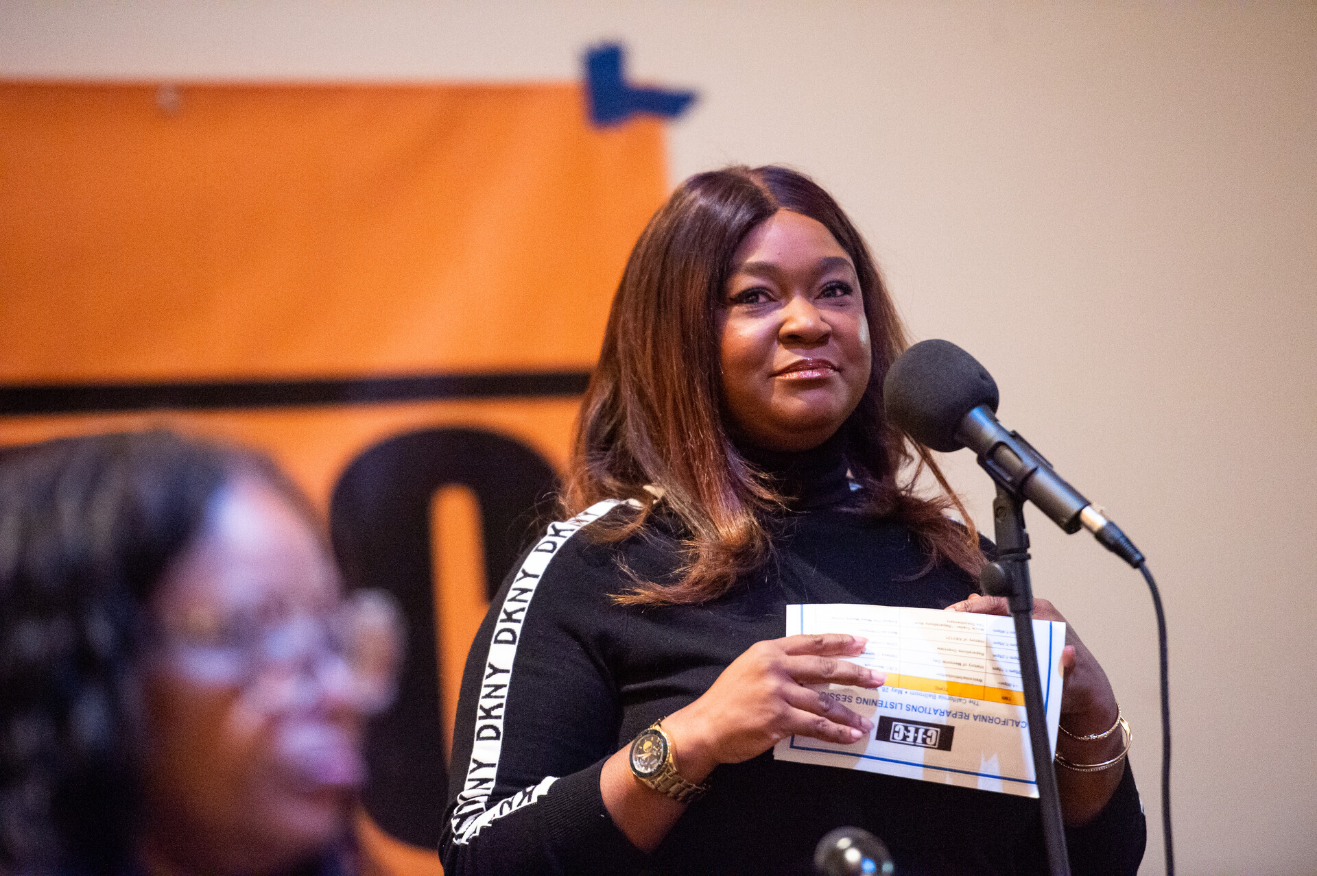 A Black woman looks towards the camera while holding a card standing in front of a microphone