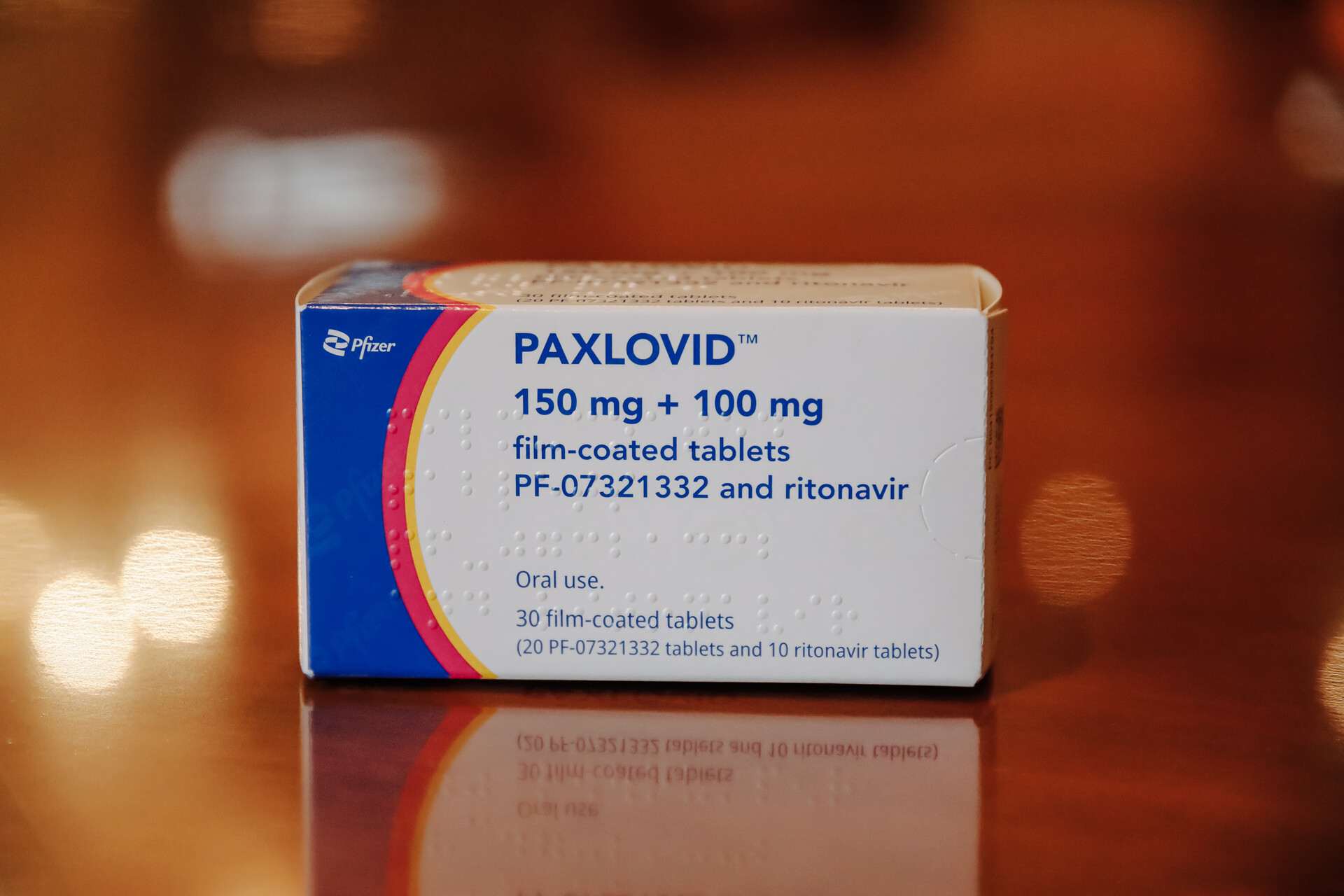A box of the COVID antiviral drug Paxlovid, placed on a shiny wooden table. The box says "PAXLOVID 150 mg + 100 mg film-coated tablets". 