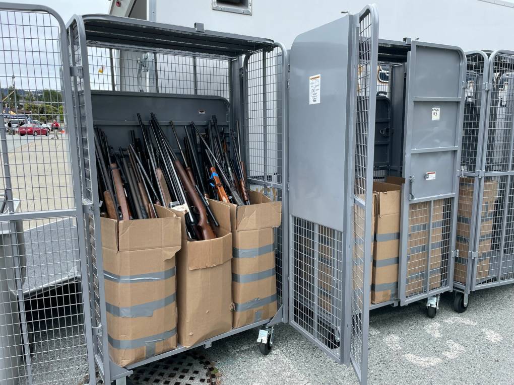 Several tall, gray, metal lockers in a parking lot hold multiple cardboard boxes full of rifles. Several cars are visible far in the background.