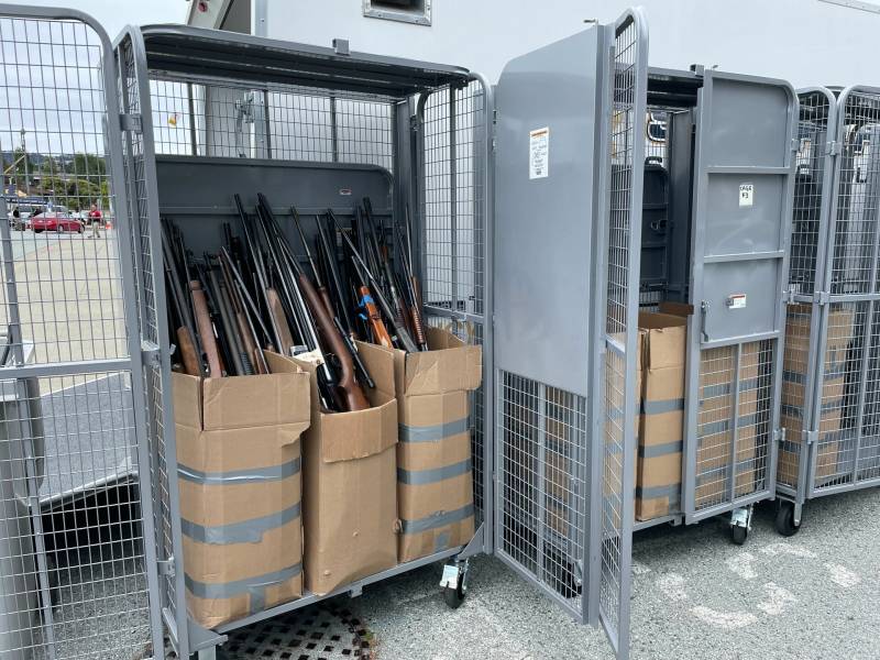 Several tall, gray, metal lockers in a parking lot hold multiple cardboard boxes full of rifles. Several cars are visible far in the background.