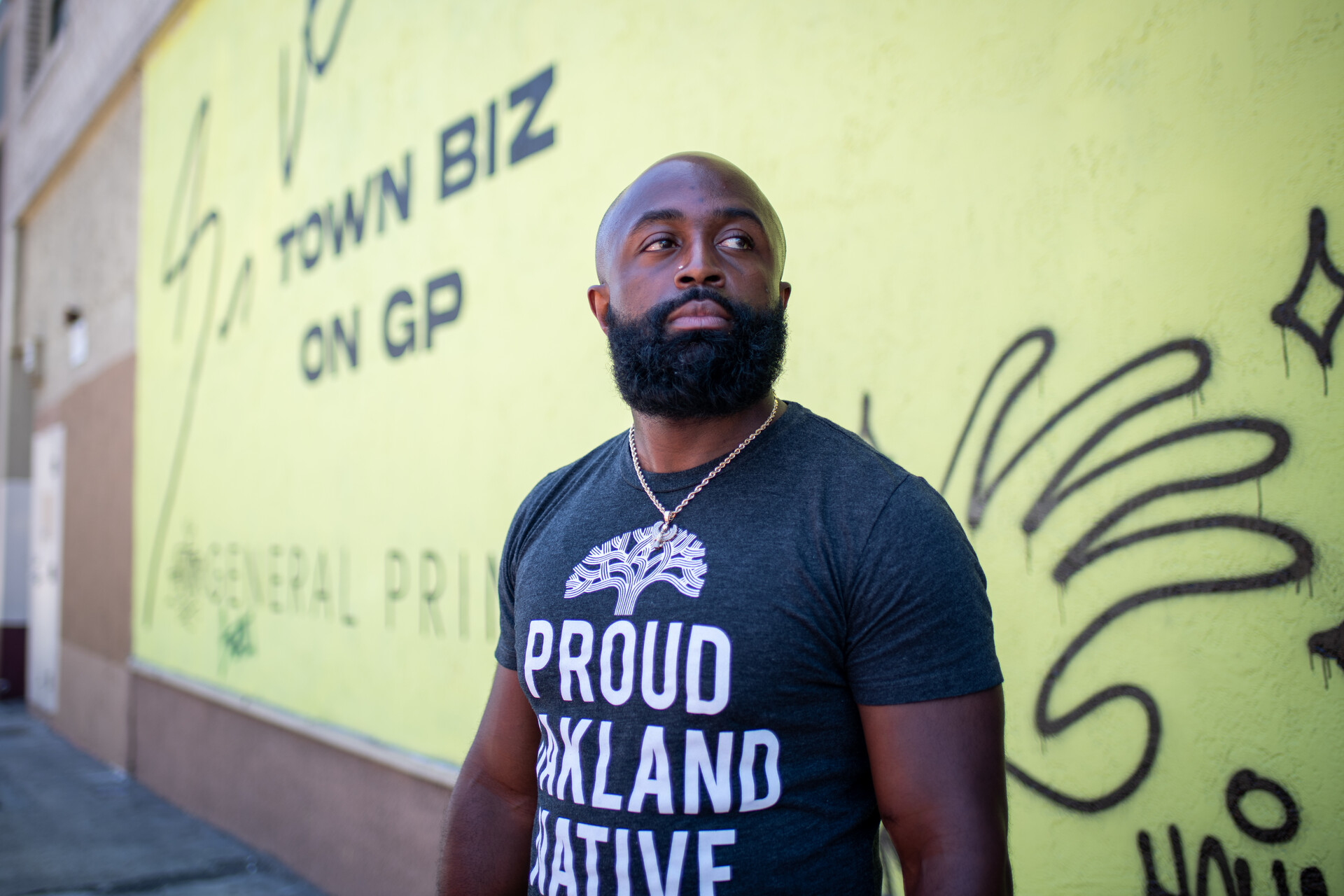  Stefon Dent sports a beard and wears his Proud Oakland Native shirt as he stars into the distance on a street in downtown Oakland. Behind him is a yellow wall with artist's tags in black paint on it.