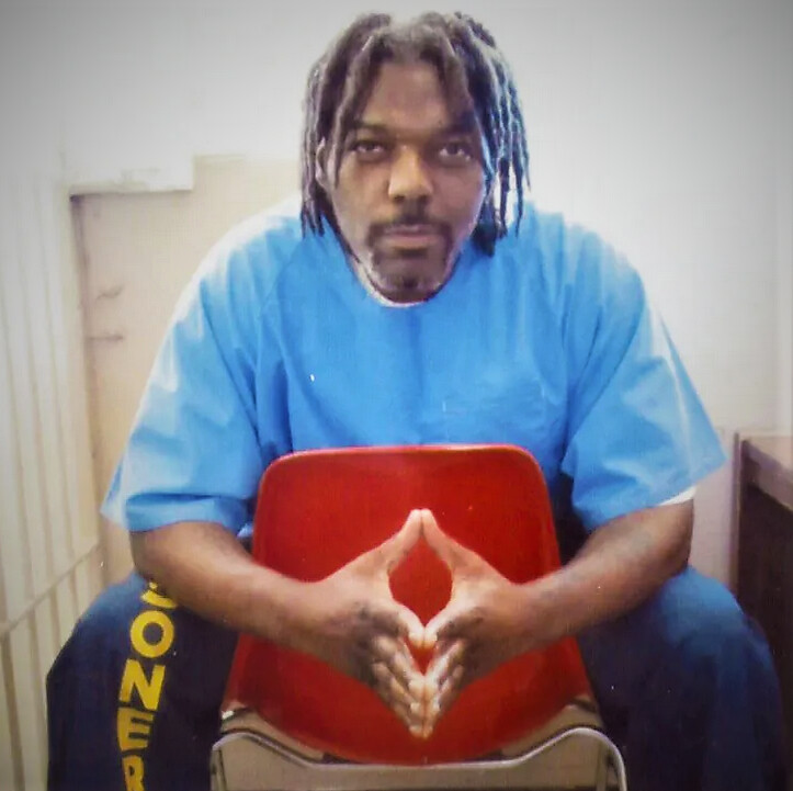 A Black man with brown eyes, chin-length locs and a graying goatee sits backward on a red plastic chair. He wears a light-blue short-sleeved prison tunic and navy blue sweatpants; on the right leg are yellow, vertical letters spelling "SONER" (as if they are part of the word "PRISONER"). He rests the fingertips of his hands, including his thumbs, together as he leans forward against the back of the chair, looking straight at the camera with a confident smile. To his right is a white-painted barred door; he appears to be inside a cell.
