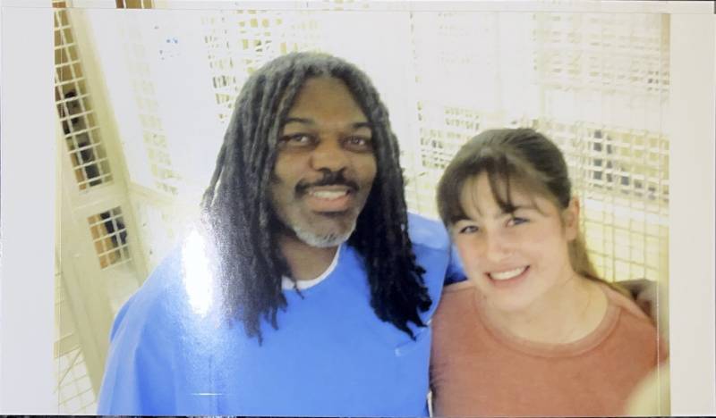A vertical glare indicates this is a picture of a photograph. A Black man with locs past his shoulders wearing a light-blue prison tunic stands shoulder-to-shoulder with a young, smiling white woman with brown bangs and a peach-colored T-shirt. They are both smiling happily.