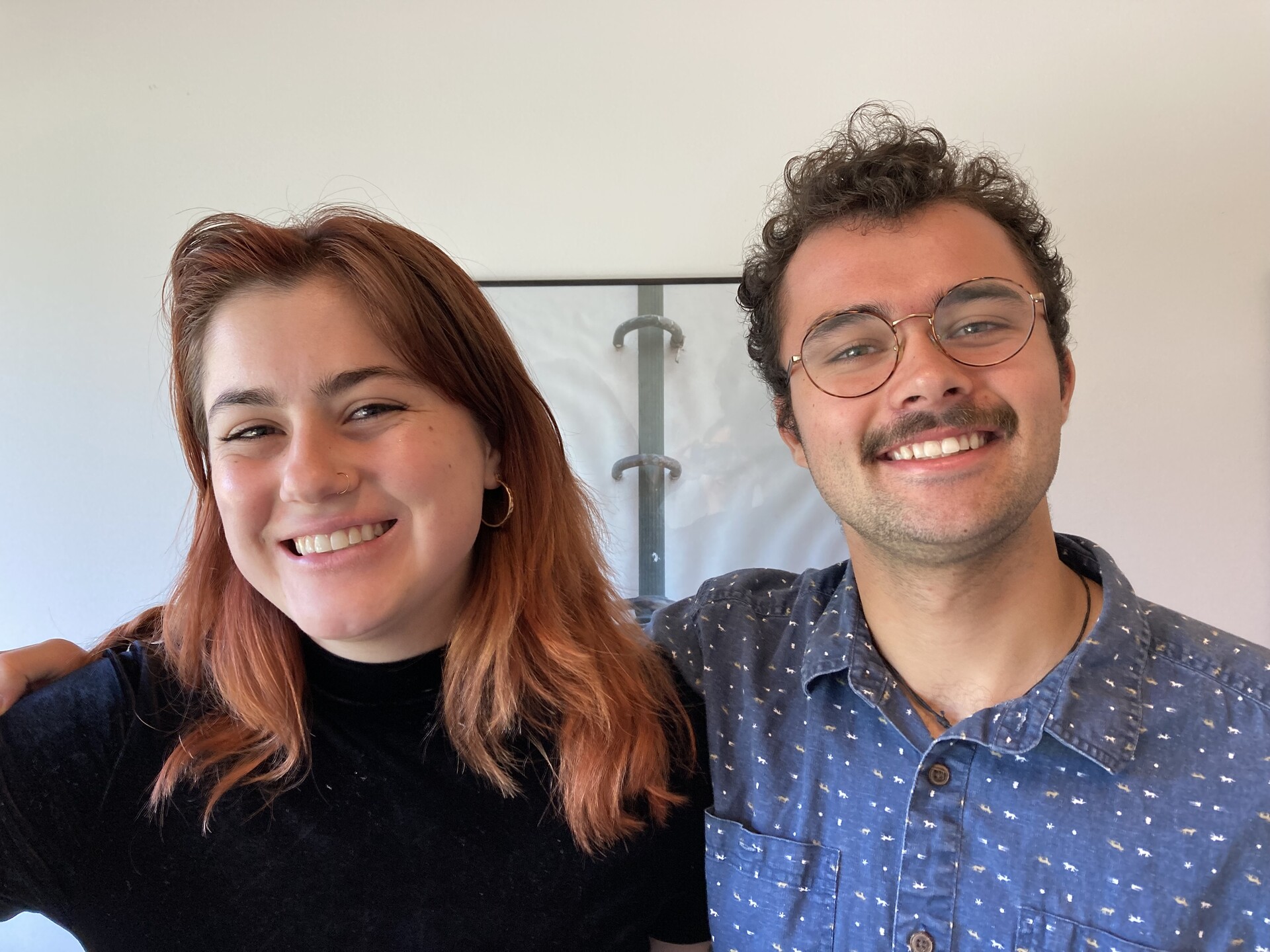 A young white woman in a black turtleneck sweater with long reddish hair smiles shoulder-to-shoulder with a young white man in a blue patterned button-down shirt, mustache and glasses, also smiling, with his right arm around her shoulders. They both look happy.