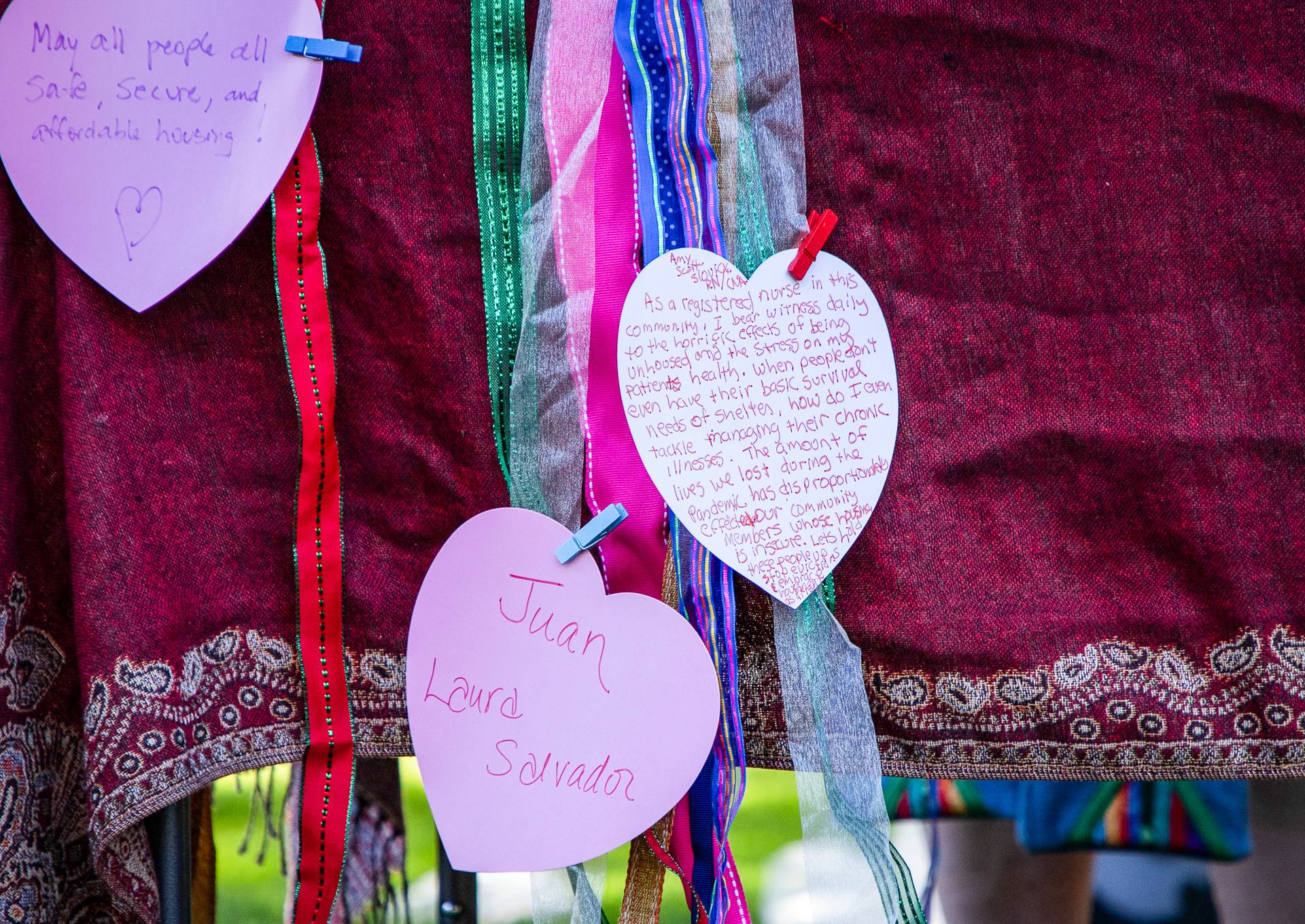 A close-up of three paper hearts -- white, pastel pink, and pastel purple -- with handwritten notes in red and purple pen, clipped with tiny blue and red wooden clothespins to multicolored ribbons: red, green, sheer, dark pink. The backdrop is a shimmery, crimson tablecloth with a paisley border, and blurry below it, on the other side of what must be a table, is a green lawn and what looks like a pair of ankles.