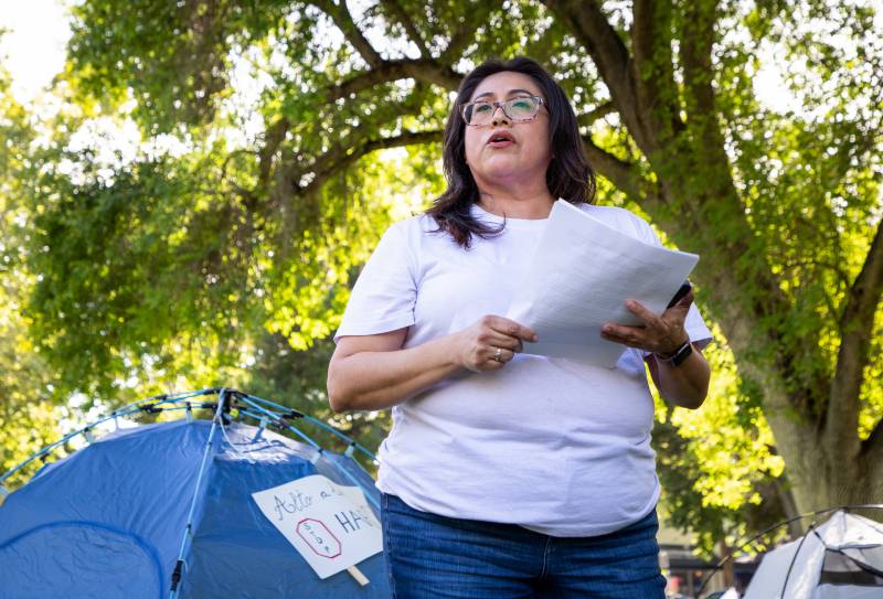 A middle-aged Latina with long, brown hair, tortoiseshell glasses, and matte red lipstick stands outside beneath a lush, green, backlit tree, wearing a white T-shirt and blue jeans, a gold ring on her right hand and a watch on her left wrist. She holds a sheaf of papers and what might be a phone in both hands as she speaks to what might be an unseen audience beyond and to the right of the camera. Behind her, to her left, is a dark blue tent with a handwritten sign on top. The words visible are "Alto a ... " on the top line and "Stop ... " on the bottom line. Behind her, to her right, is the top of a second, white tent. Barely visible in the background is what looks like a storefront, indicating this might be in a city park.