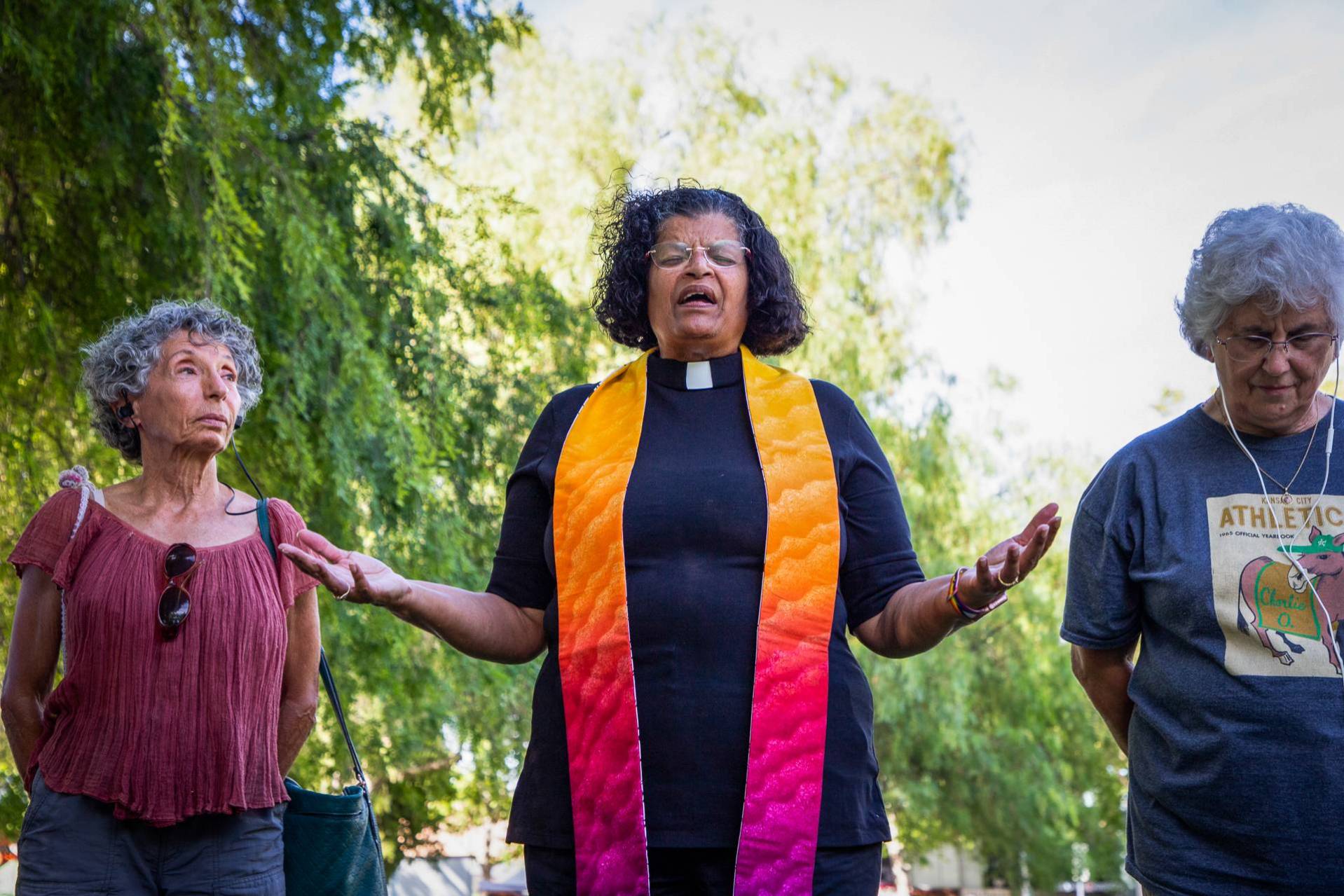 Three people stand in a row outdoors before a backdrop of lush, green trees. The woman in the center, a middle-aged Latina with curly, shoulder-length black hair and wireless glasses, wears a short-sleeved black cassock with a white clerical collar, and a bright stole of a warm yellow that turns to orange, then pink, then purple on both sides. She has her eyes closed and her arms held out, palms up, and appears to be speaking. On both sides of her stands a middle-aged women, one with short gray hair, the other with white gray hair. Both are dressed casually in short-sleeved tops. The woman to her right stands slightly behind her, looking at her as she speaks, sunglasses tucked into her rose T-shirt and a purse hanging from one shoulder. The woman on her left bows her head, hands behind her back, the white wires of headphones coming from both ears.