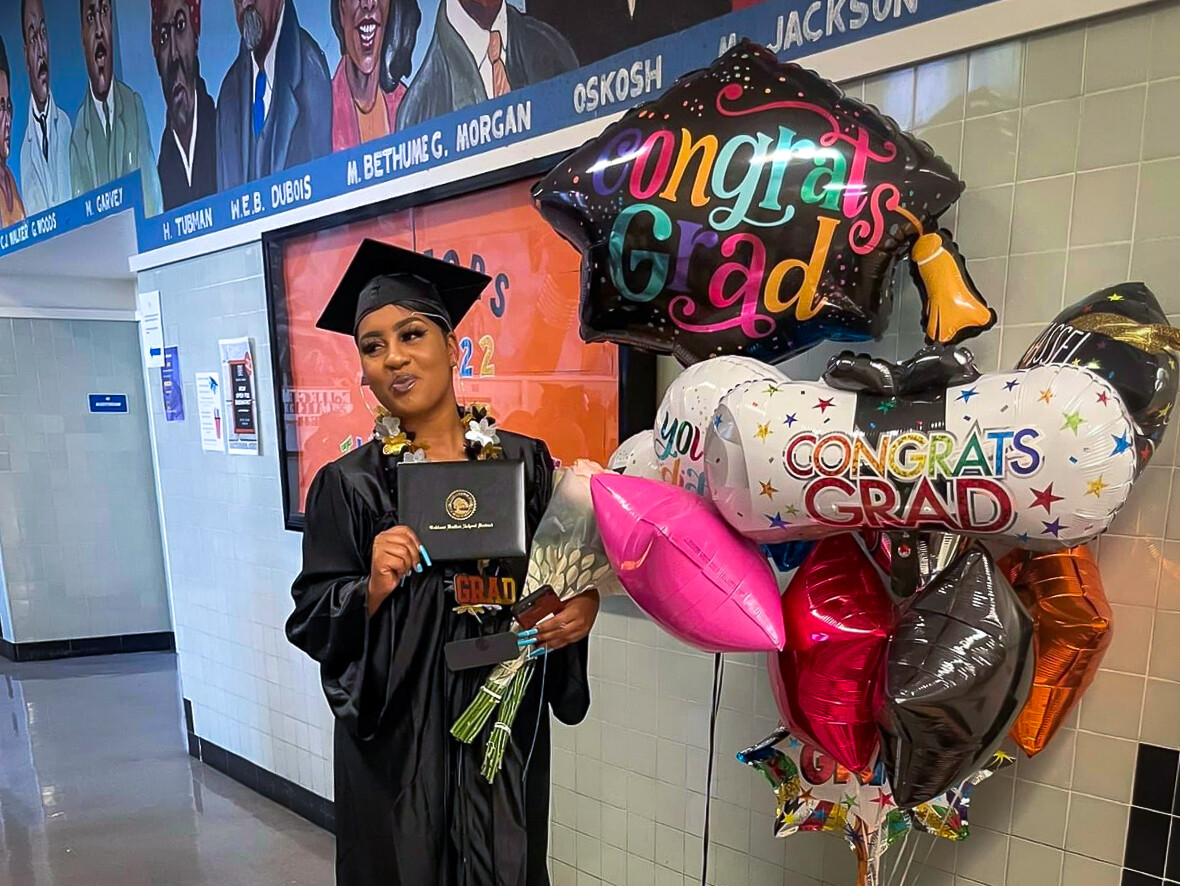A woman stands in a school hallway next to graduation balloons. She is holding up a diploma and is smiling at someone off-camera.