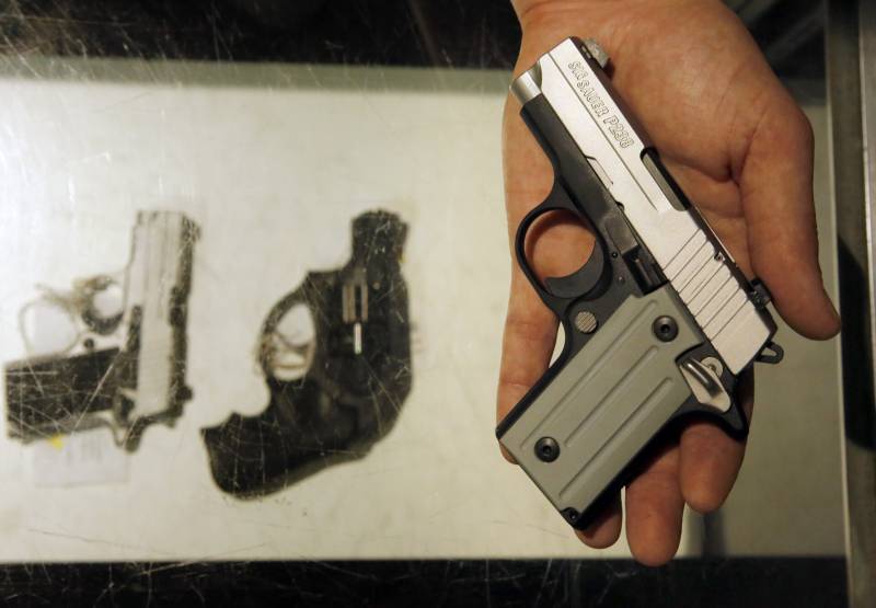 hand holds small gun above glass case containing two other small handguns