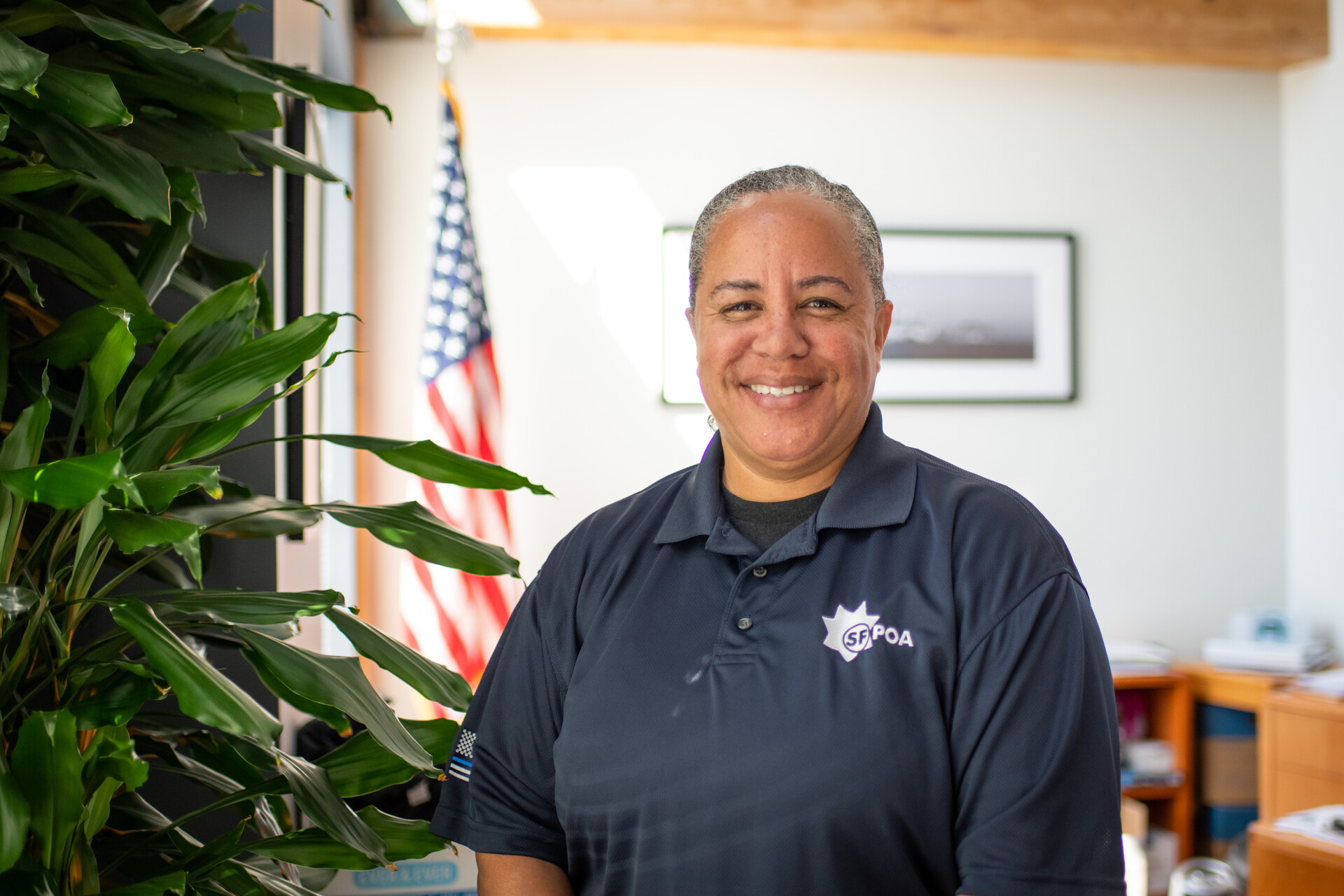 A Black woman smiles broadly, wearing a blue SFPOA shirt in an office with a US flag in the background