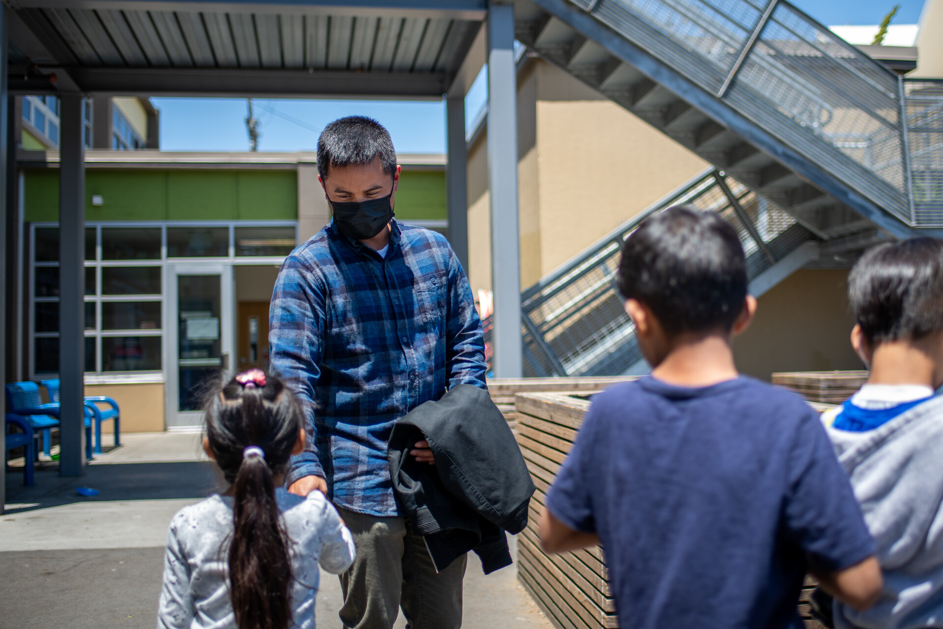 A teacher wearing a mask speaks with young students outside. 