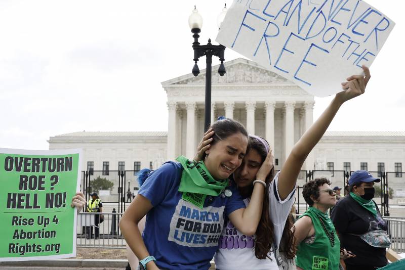 two women hold protest signs reading 'never land of the free' while they hold each other and cry in front of the supreme court building