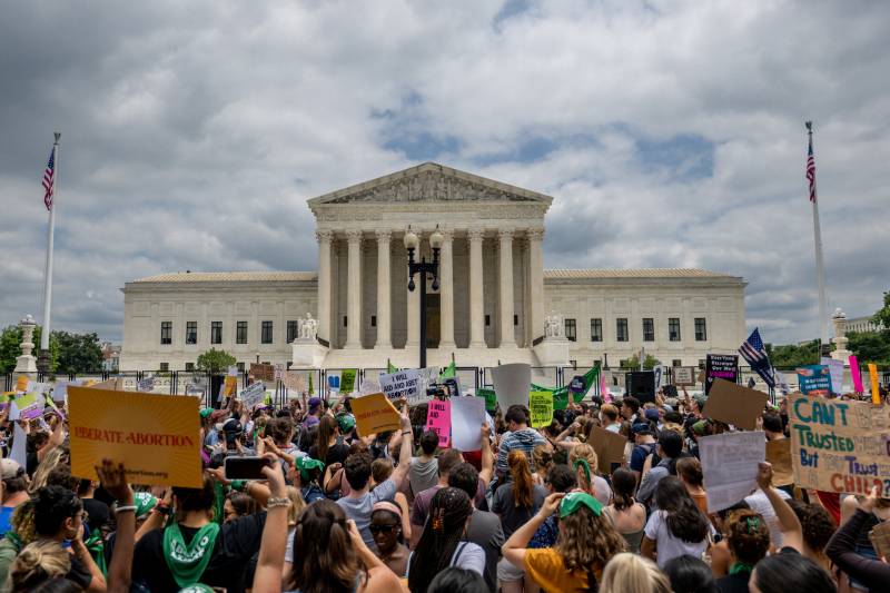 The front of the U.S. Supreme Court, under a dark grey and blue cloud sky, with a crowd of people protesting outside and holding up signs.