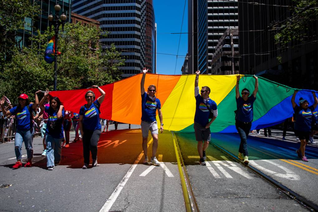 Several people are seen walking down the middle of the street holding up a large rainbow flag.