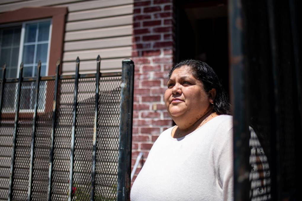 Sulima Navarrete, who is Latina, stands outside her home wearing a white shirt. Her hair is dark brown and pulled back. She is standing in a doorway, by a black metal fence with brickwall behind it. She is unsmiling.