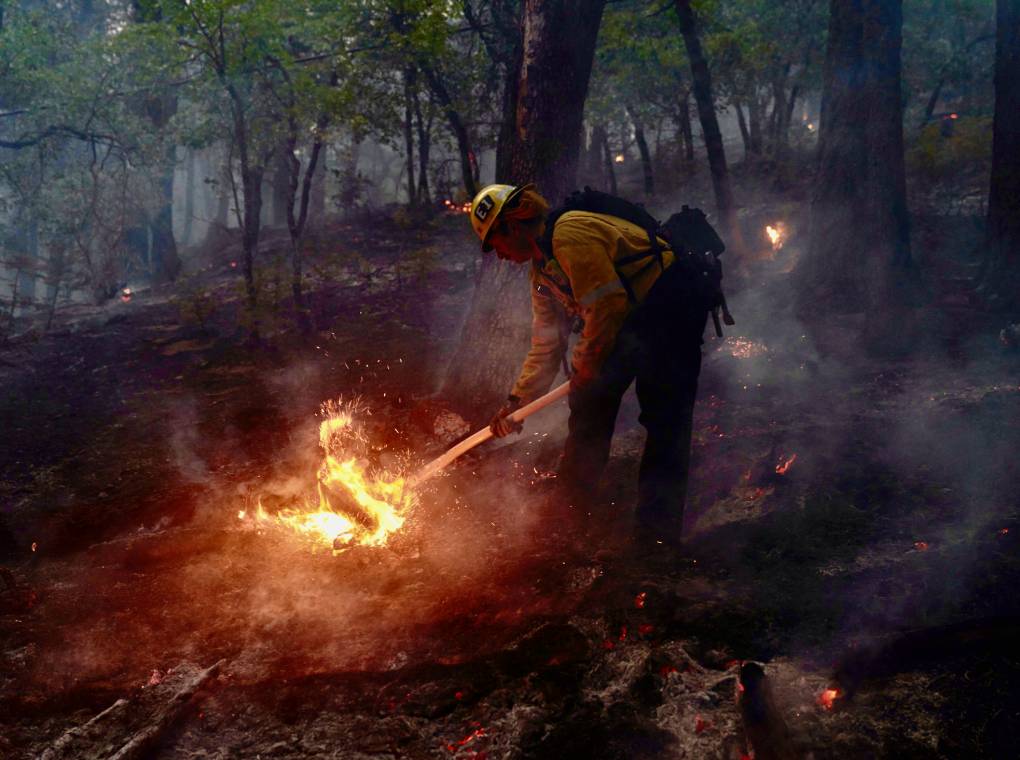 A man wearing a yellow helmet and firefighter suit uses a long object to put out a fire.