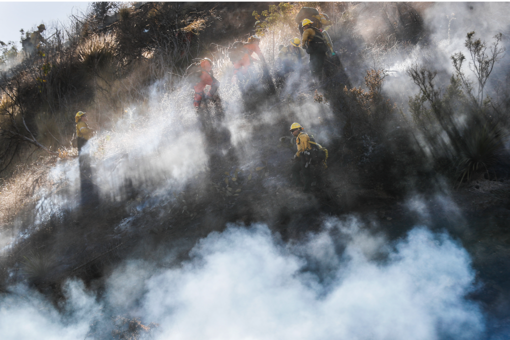 Entire frame, except top-left corner, is a brown hillside, partially obscured by smoke, which is lit by rays of sunlight from above. The top-left corner of the frame confirms it's a clear day. About eight wildland firefighters dressed either in all-orange or all-yellow gear, including helmets, appear to be digging into the brown dirt and brush.