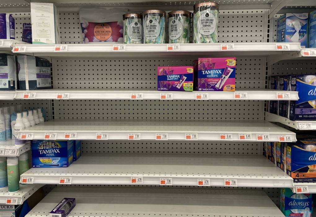 Five white metal almost completely empty shelves, probably in a pharmacy or grocery store. The bottom shelf is completely empty; the next up has a single item on it, on the far left (a black box of Astroglide lubricant); the next up has two blue boxes of Tampax brand tampons to the far left; the next shelf up is empty; the next shelf up has two rows of pink boxes of different Tampax brand tampons, on the middle right; and the top row is half full, with the most items: four rows of plastic-wrapped, cylindrical containers of "100% organic cotton care tampons," which are to the right of a single container of "ultra-thin liners," also an organic brand, at the very back of the shelf, which is to the right of a single, cream-colored box of something turned to the side. Peeking out to the left and right of this set of shelves are obviously full shelves of (to the left) shampoo products and (to the right) feminine pads (Always brand).