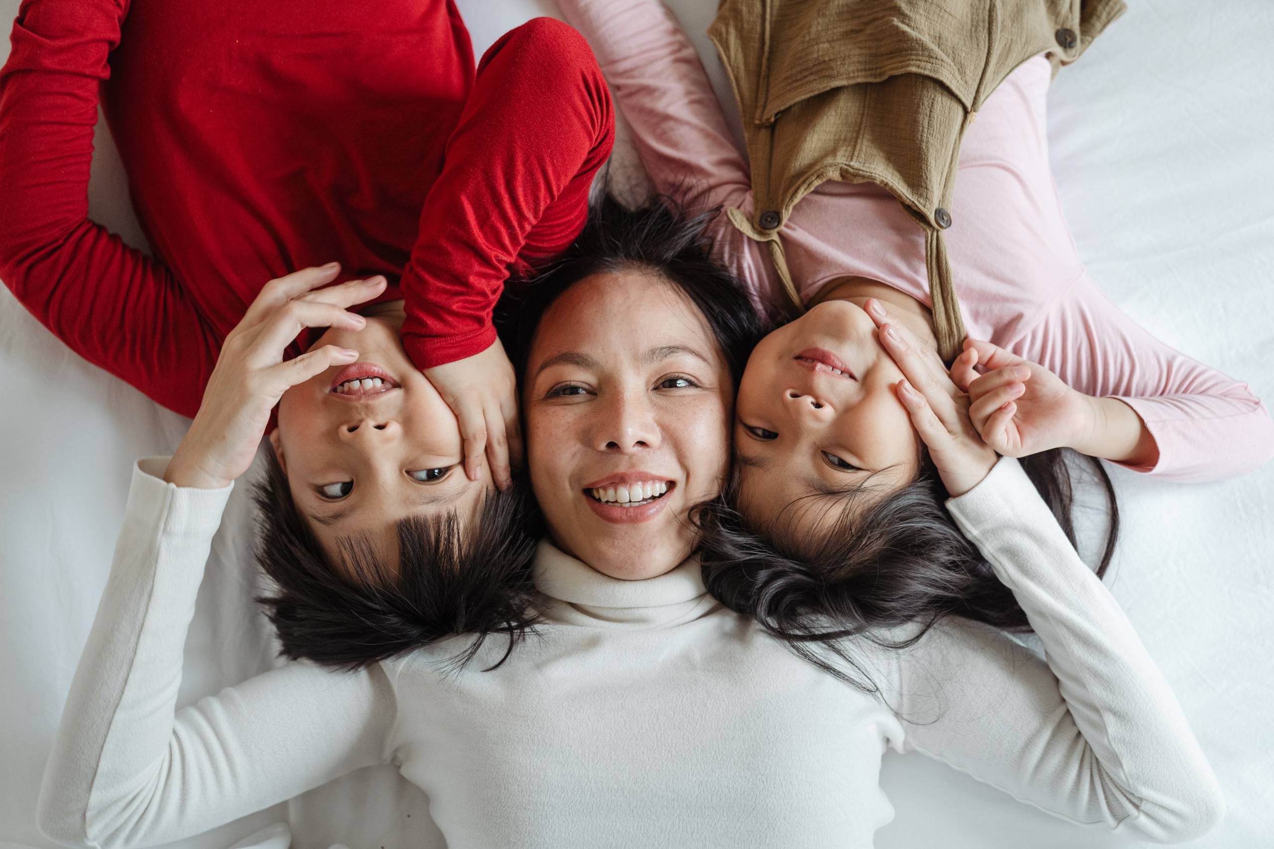 A woman wearing a white turtleneck is lying on a bed with two children on either side of her. One of them is a boy wearing a red shirt, and to her right is a girl wearing a light pink shirt and brown overalls.