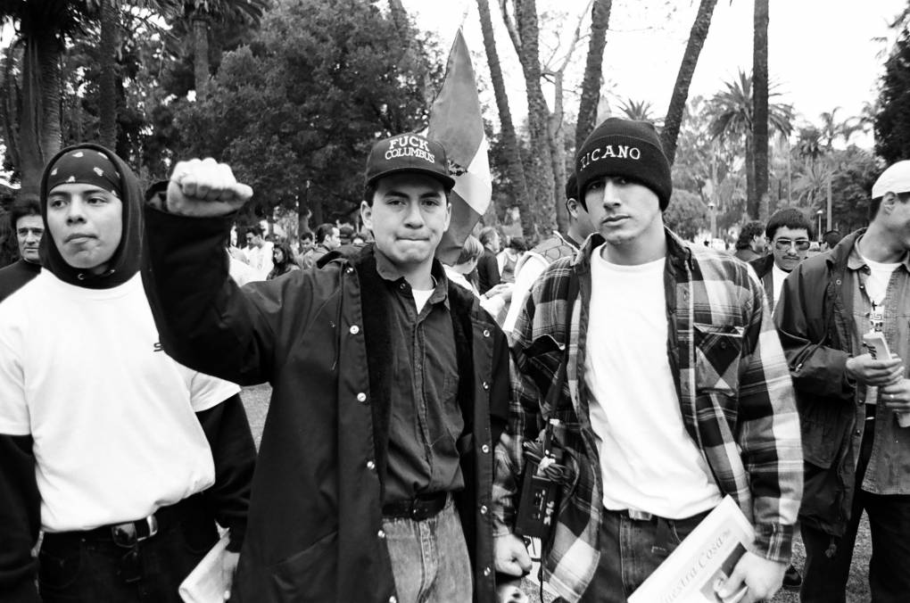 black and white photo showing two young men at a rally, one of them raising his fist in solidarity, wearing a black cap reading 'fuck Columbus'