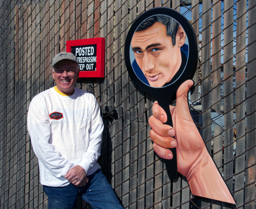 A middle aged man in a white long sleeved tee shirt and baseball hat poses near a fence. In a trick of perspective, it looks like a giant hand next to him holds a mirror with James' Dean's face reflected.
