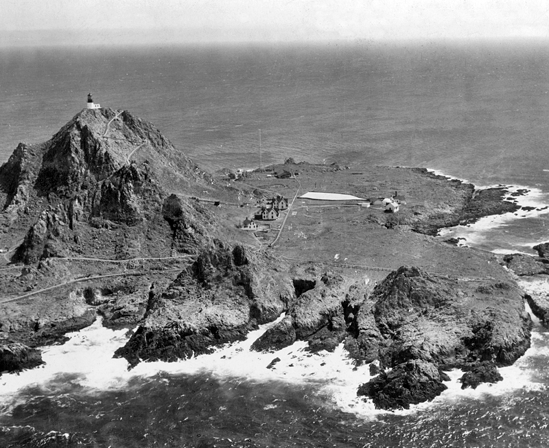 A black and white 1929 photograph of the Southeast Farallon from the sky. You can see three dwellings, a lighthouse, and several auxiliary buildings. 