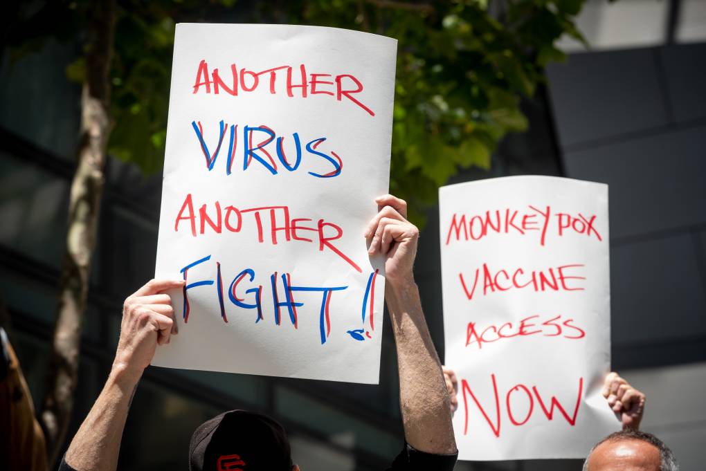 Two people hold signs at an outdoor protest. One sign says, 'Another virus another fight'. The second one says, 'Monkeypox vaccine access now'.