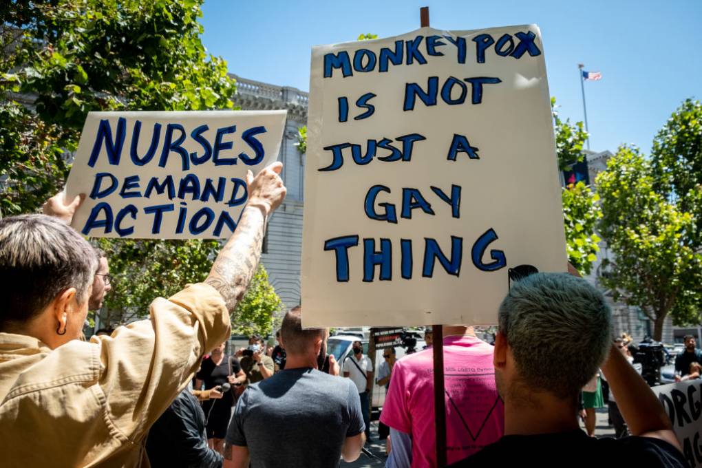 Demonstrators standing outside a large building holds signs that say 'Nurses demand action' and 'Monkeypox is not just a gay thing'.