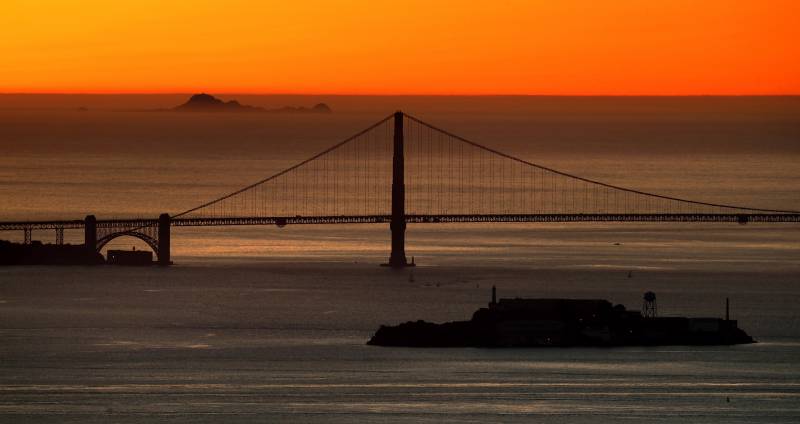 The sun setting over the Pacific ocean. The Golden Gate Bridge is an silhouette, and on the horizon, the Farallon Islands stick out of the ocean. 