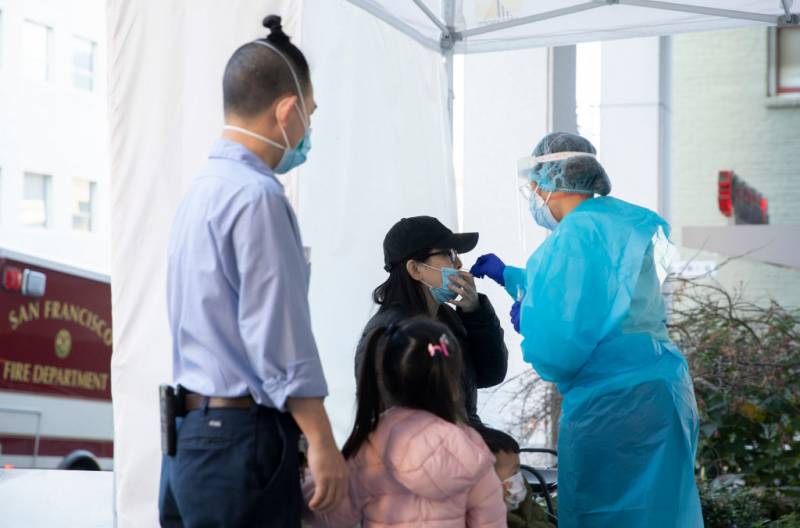 A Chinese woman (her ethnicity confirmed by the reporter) wearing a black jacket is having her nose swabbed as part of a COVID test by a medical worker to her right, who is wearing bright blue scrubs and a hair protector. A man in a blue shirt and a child in a pink jacket are in the foreground, watching the swab happening.