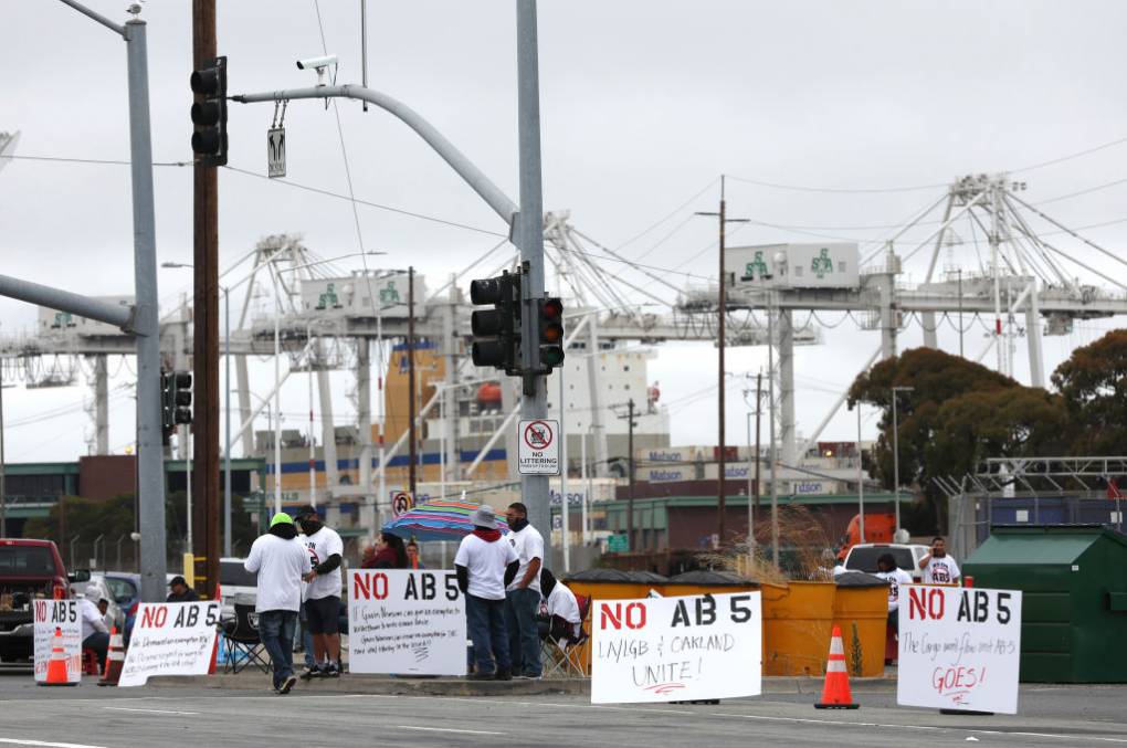 A group of workers stand at a port, large cranes in the distance, next to signs that say 'No AB 5.'
