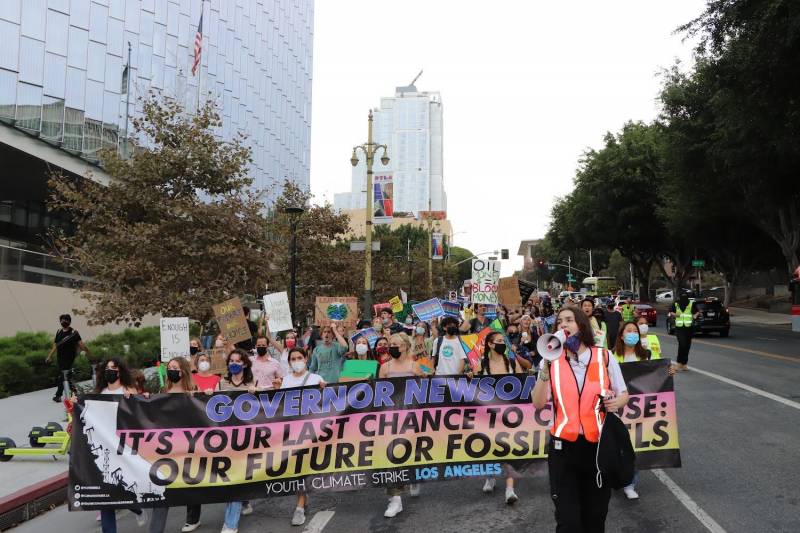 Young people carry a banner in a climate march reading, "Governor Newsom: It's your last chance to choose: Our Future or Fossil Fuels. Behind the banner is a crowd of young people wearing covid masks and carrying signs. At the front, Paola Hoffman speaks through a bullhorn. There are green trees on either side of the road and a glassy building on the left with an American flag in front of it.