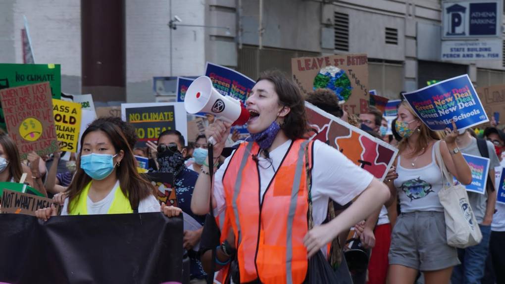 A woman with white skin and dark hair speaks loudly through a bullhorn. She's wearing a white t-shirt and an orange traffic safety vest. Young people holding signs supporting climate action march to the sides and behind her. The signs read "Green New Deal" and "No Climate No Deal." The young people are of multiple races and wearing covid protection masks.